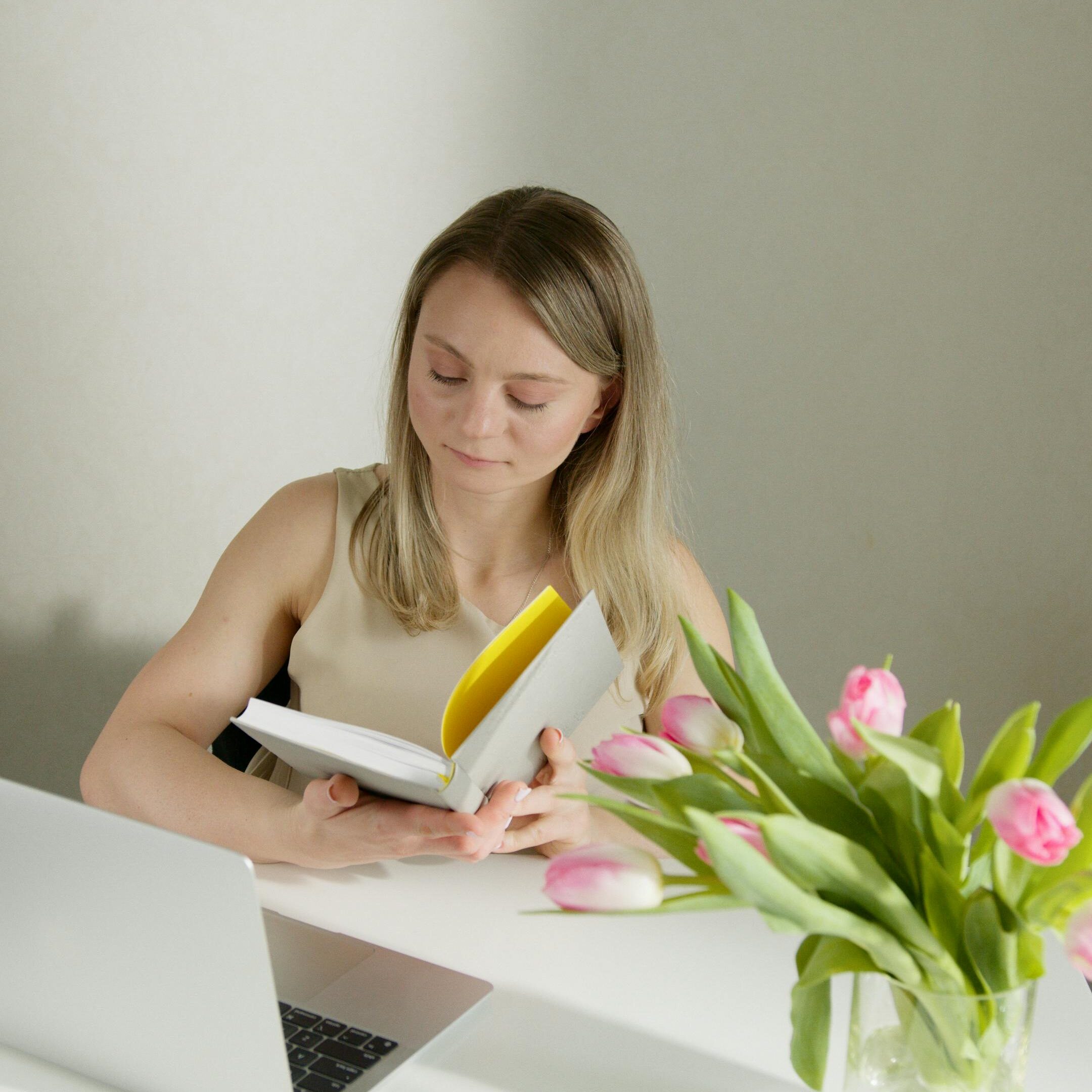 A woman reading a book at a bright desk, accompanied by a laptop and fresh tulips in a vase.